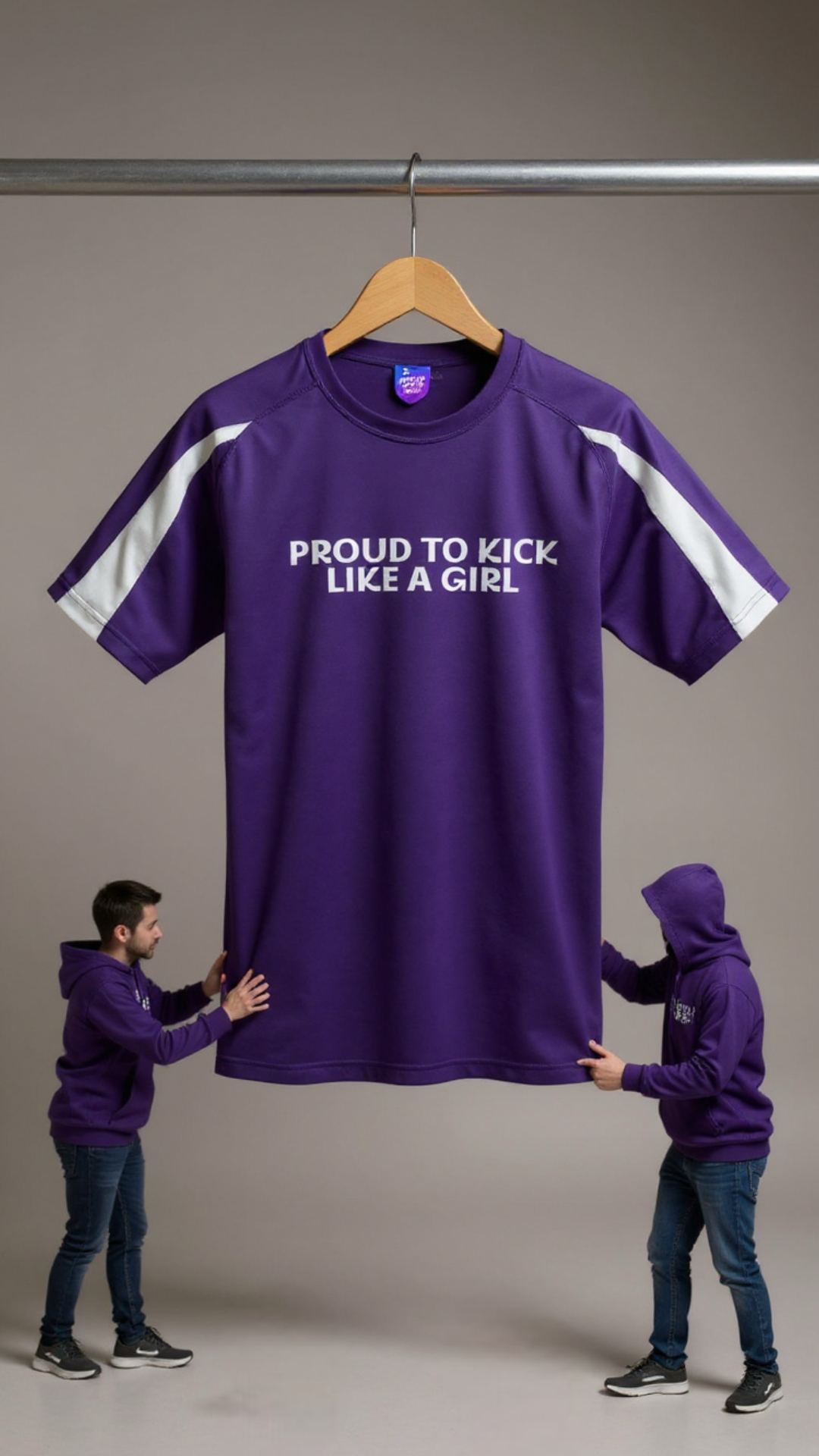 Purple t-shirt with 'PROUD TO KICK LIKE A GIRL' text on a hanger, with two people in matching purple hoodies holding it.