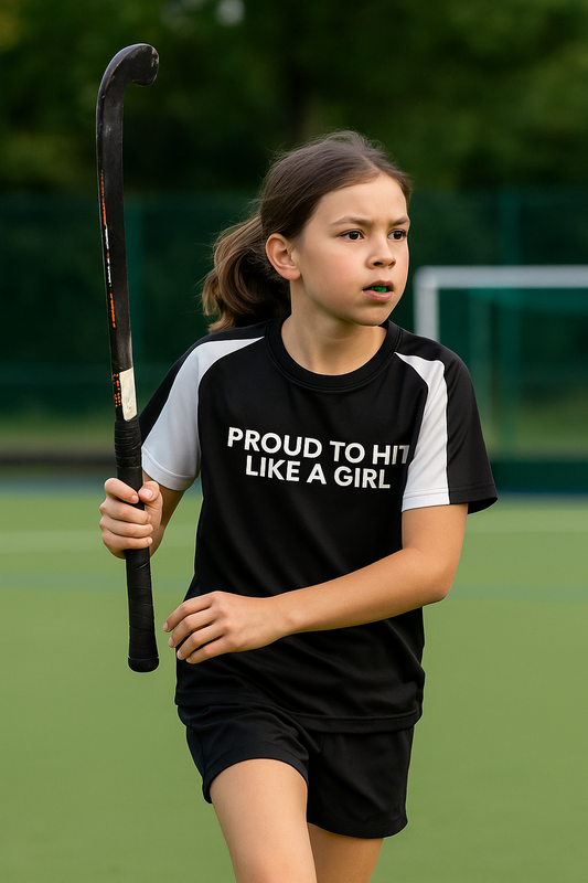Young girl holding a hockey stick on a sports field wearing a shirt with 'PROUD TO HIT LIKE A GIRL' text.