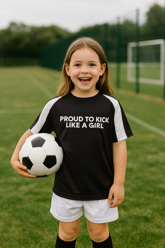 Young girl holding a soccer ball on a sports field wearing a black t-shirt with 'PROUD TO KICK LIKE A GIRL' text.